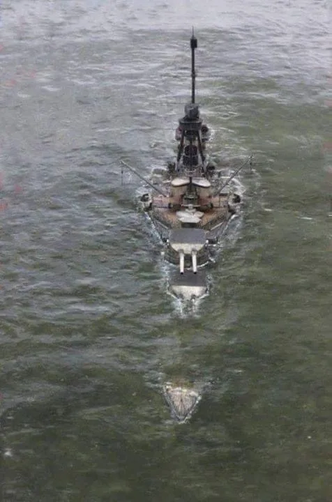 An aerial view of a large battleship moving through choppy greenish water, with its bow pointed forward and a visible wake…