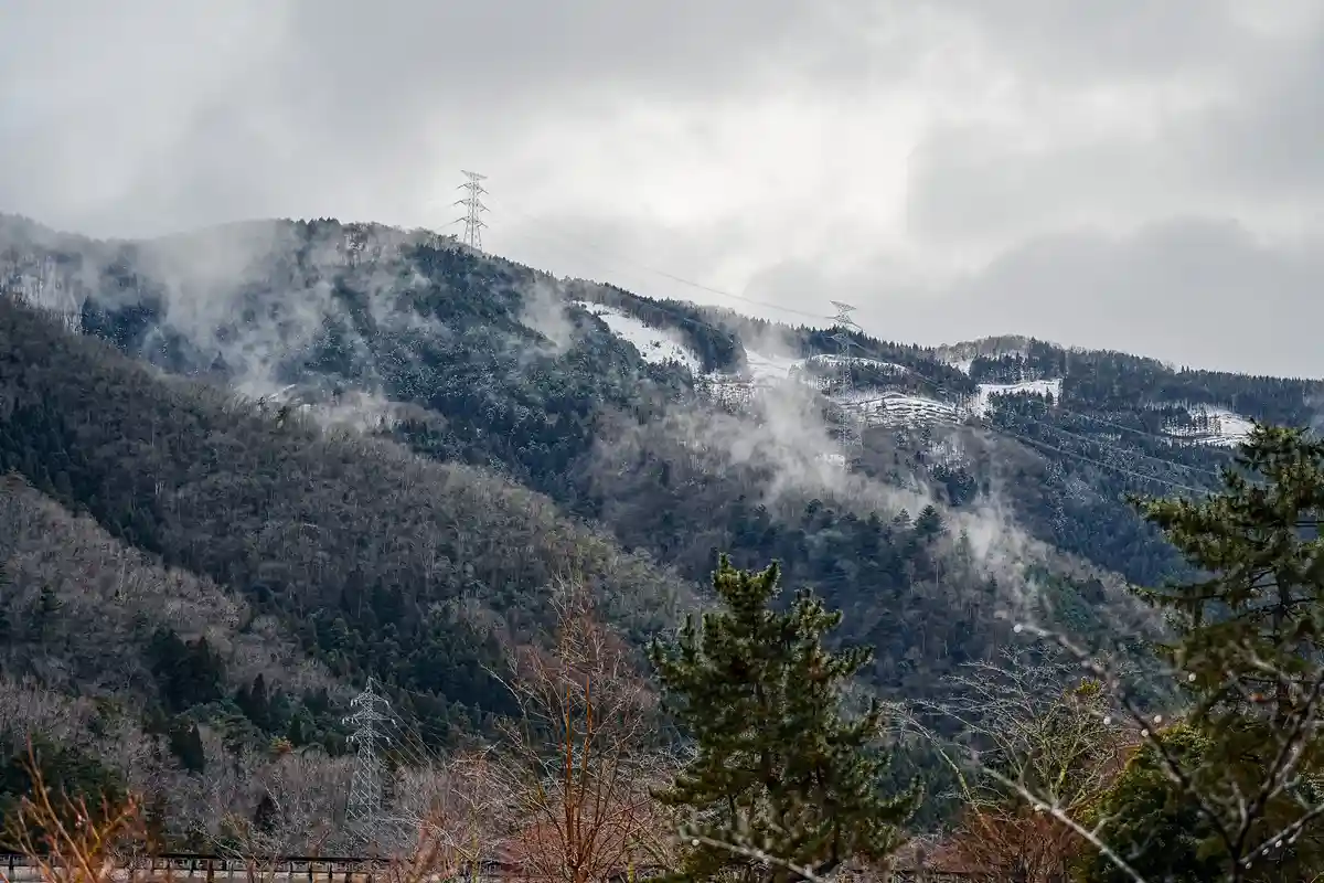 A misty, snowy mountain landscape with evergreen and bare trees, power lines and towers crisscrossing the hills under a…