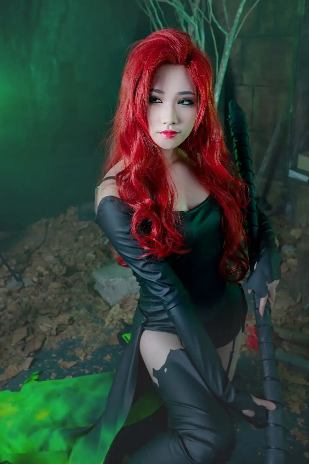 A person in a Poison Ivy cosplay with long red hair, green eyes, and a black leather outfit, holding a staff in a dark…