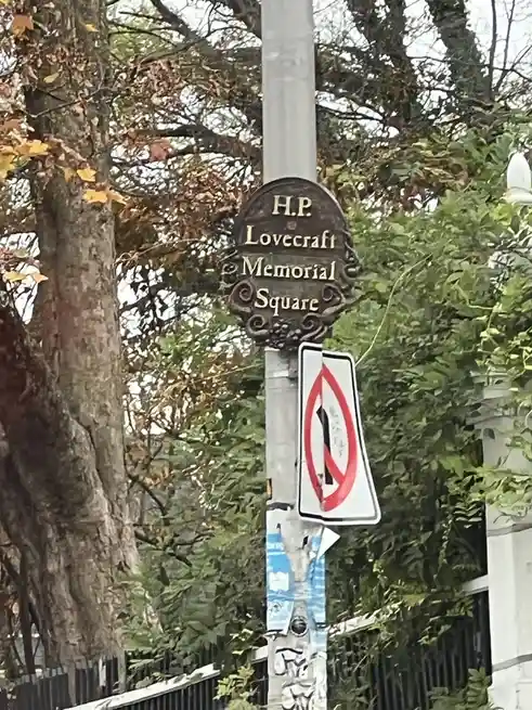 A street pole displays a bronze plaque reading 'H.P. Lovecraft Memorial Square' above a standard 'no left turn' traffic…