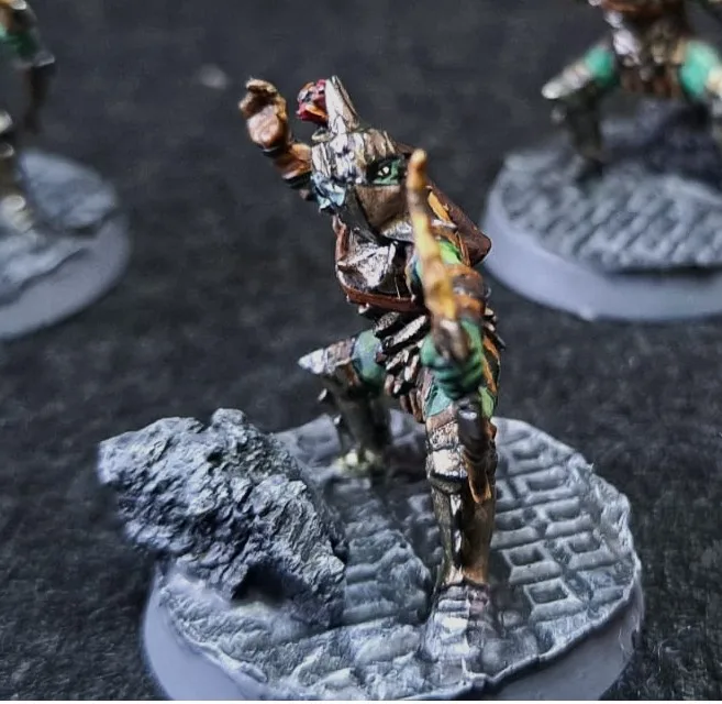 A painted miniature figurine of a fantasy warrior with a bird-like head, standing on a stone base with a rock beside it. The…