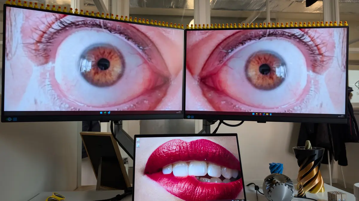 Two large monitors display extreme close-ups of human eyes, with a row of yellow rubber ducks lined up along their top edge…