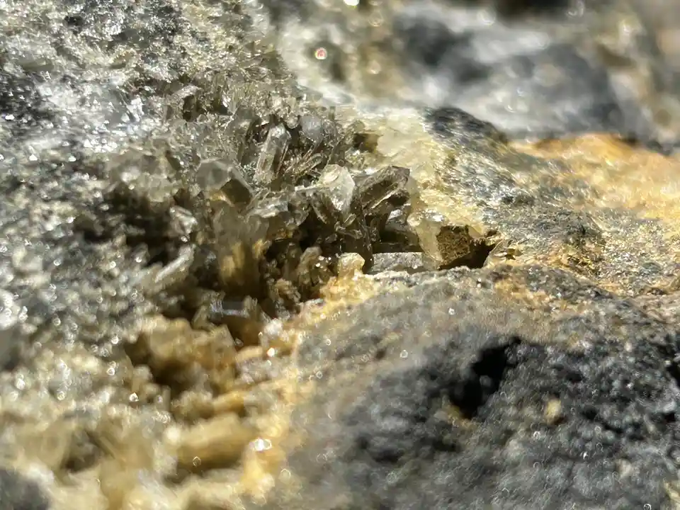 A close-up photo shows a cluster of small, translucent, glassy crystals growing out of a rough, textured rock surface with…
