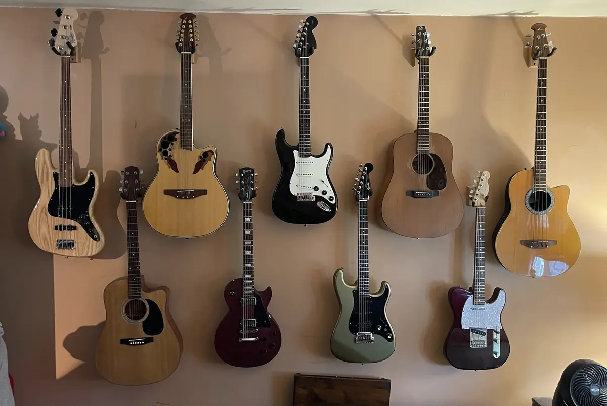 A wall displays ten guitars of various types and colors, including electric and acoustic models, hanging in two rows. The…