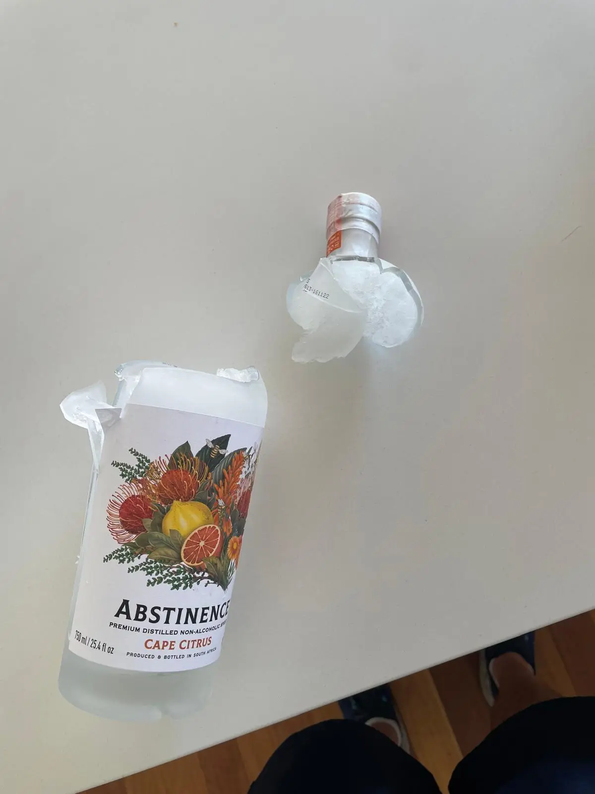 Two bottles of 'Abstinence' non-alcoholic spirit sit on a white table, both visibly crushed and broken. The larger bottle on…