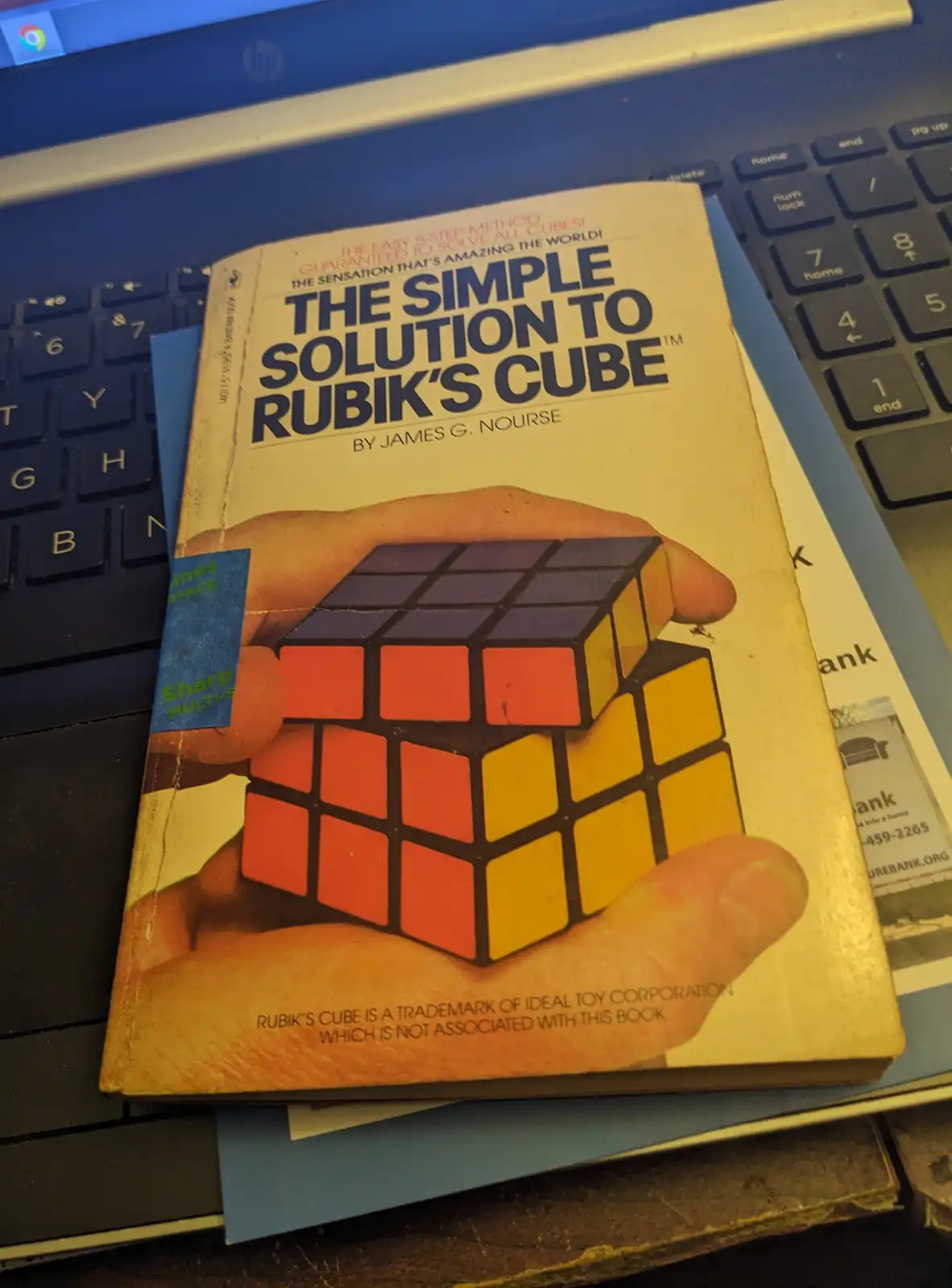 A vintage paperback book titled 'The Simple Solution to Rubik's Cube' by James G. Nourse rests on a laptop keyboard. The…