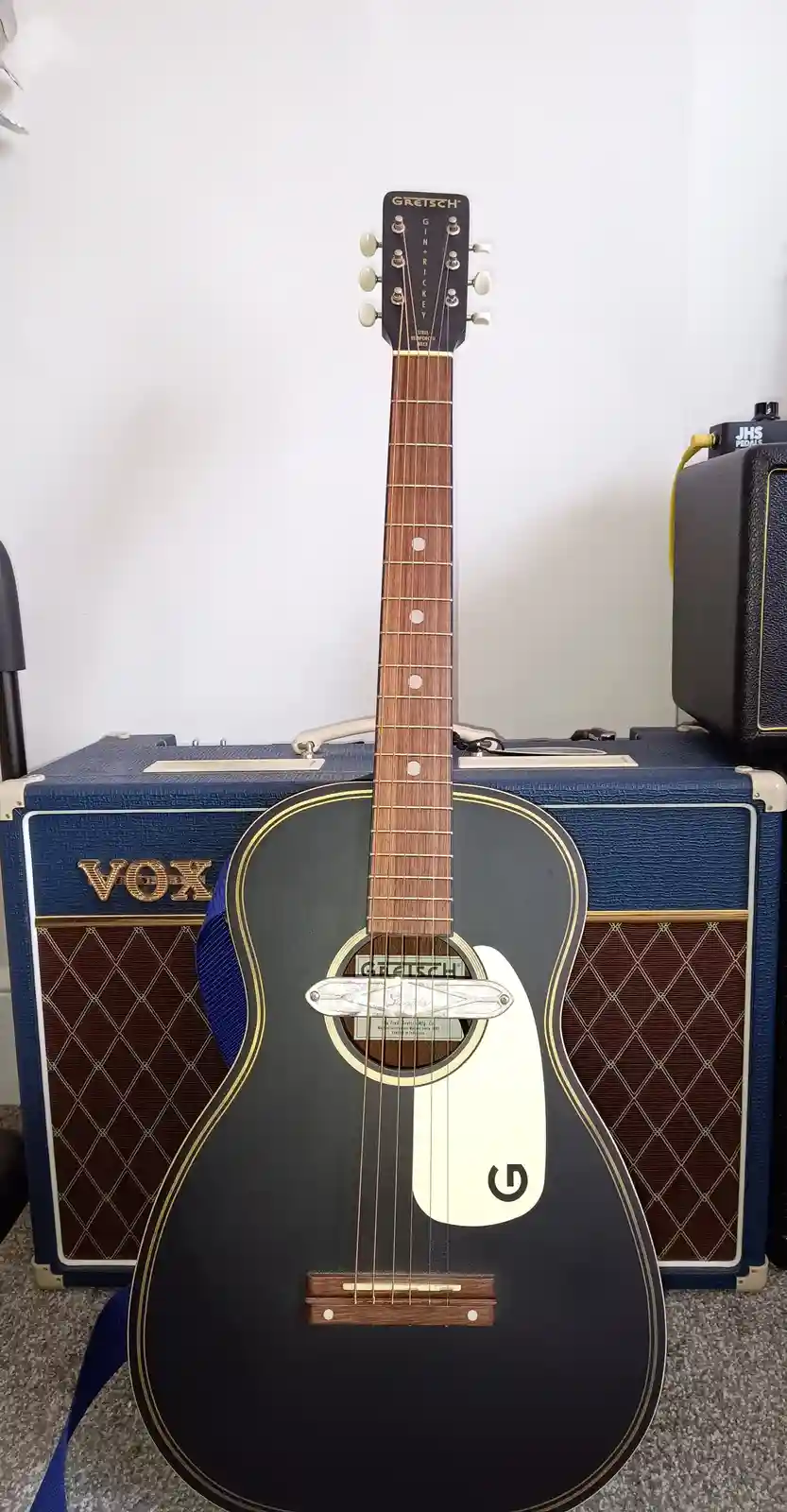 A black Gretsch acoustic guitar with a white pickguard and gold trim stands upright in front of a blue Vox amplifier. The…