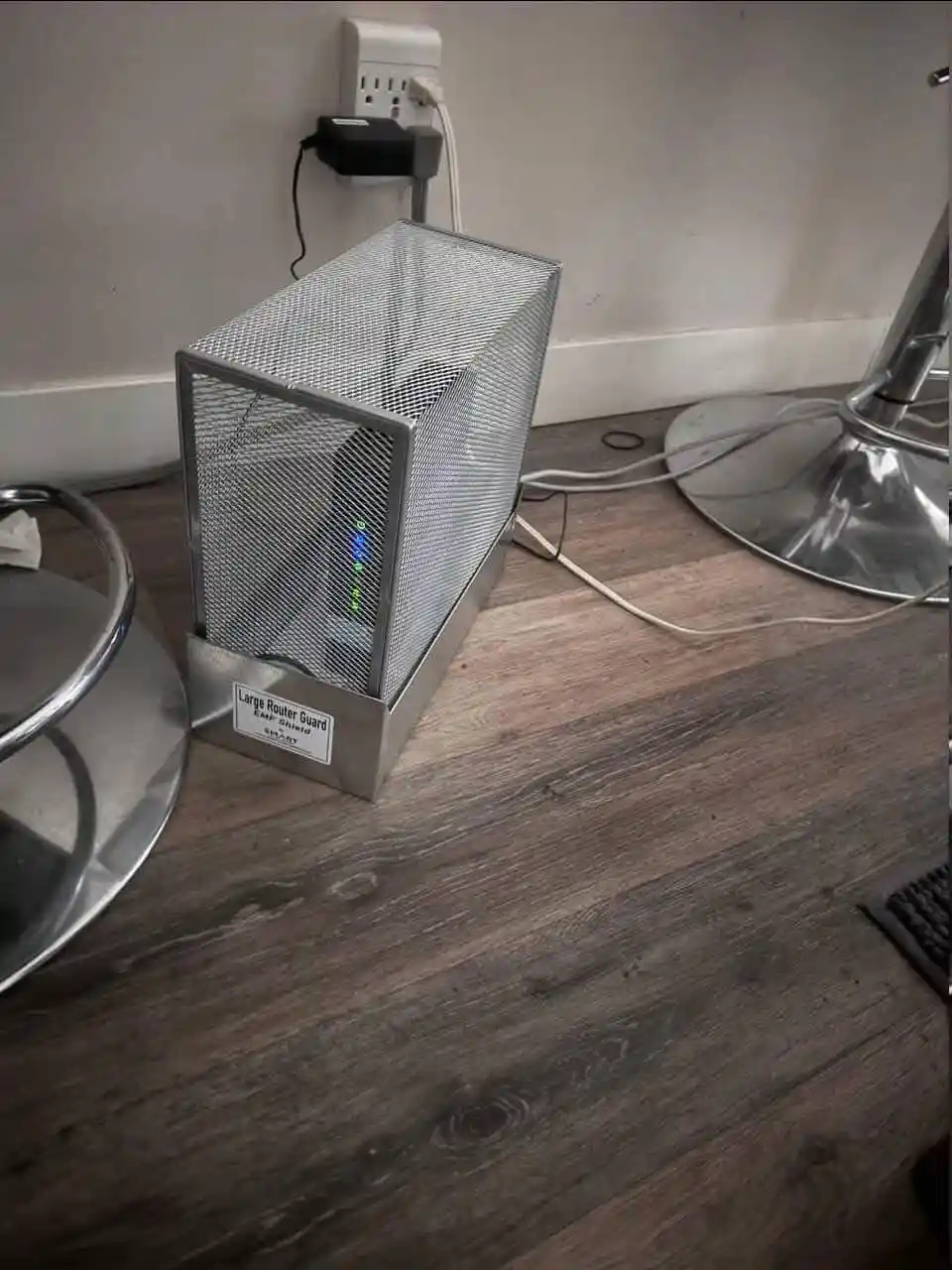 A router sits inside a large, silver mesh metal container labeled 'Large Router Guard EMF Shield' on a wooden floor, with…