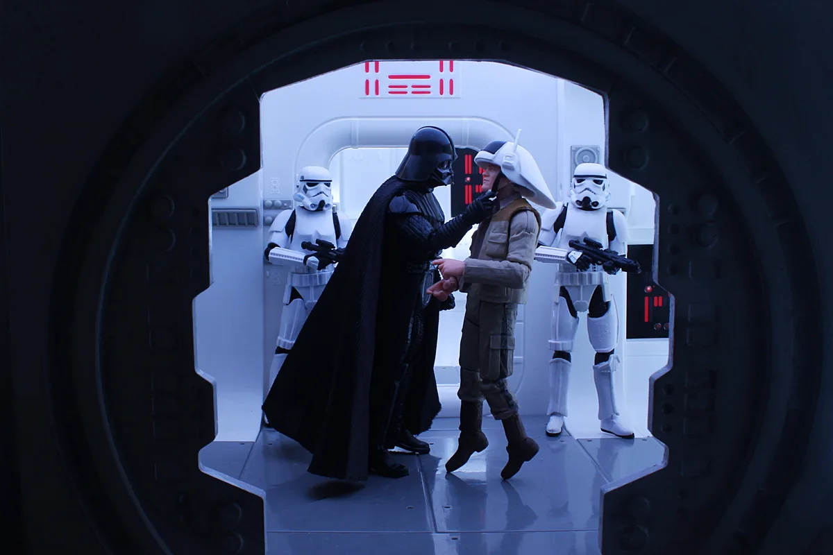 A photo of Star Wars action figures: Darth Vader in black armor and cape confronts a Rebel soldier in tan gear, with two…