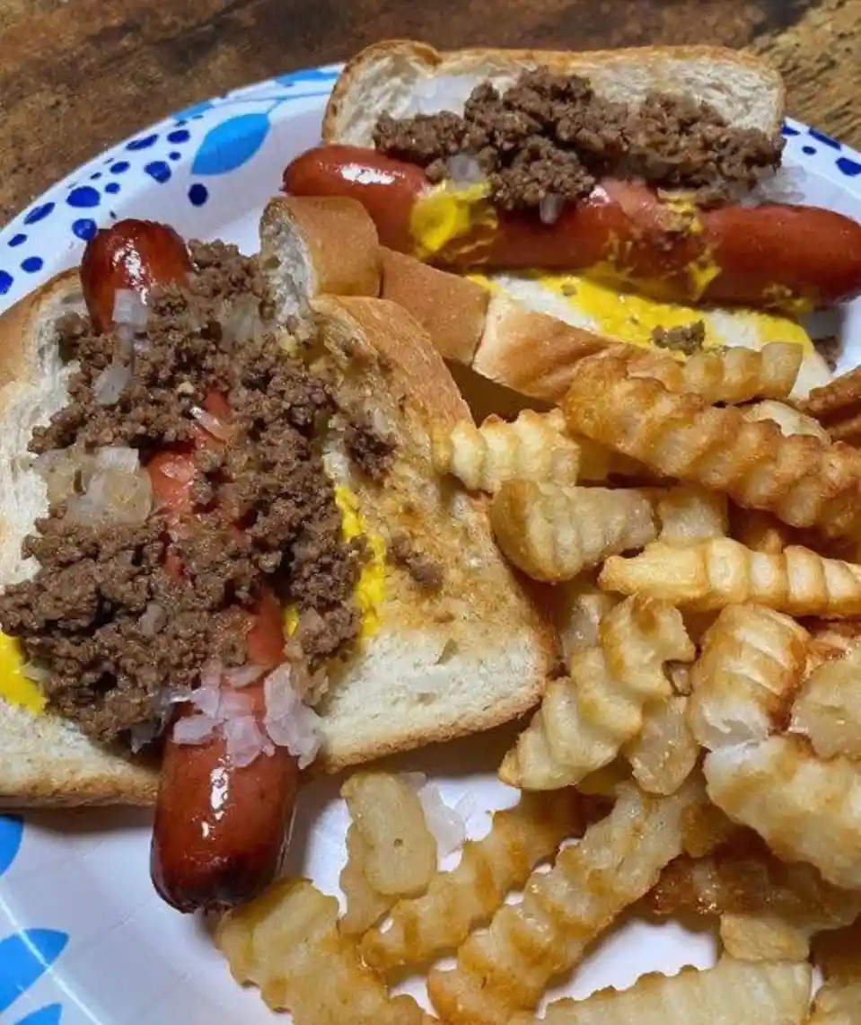 A close-up photo of two hot dogs in buns, generously topped with chili, onions, and mustard, served alongside crinkle-cut…