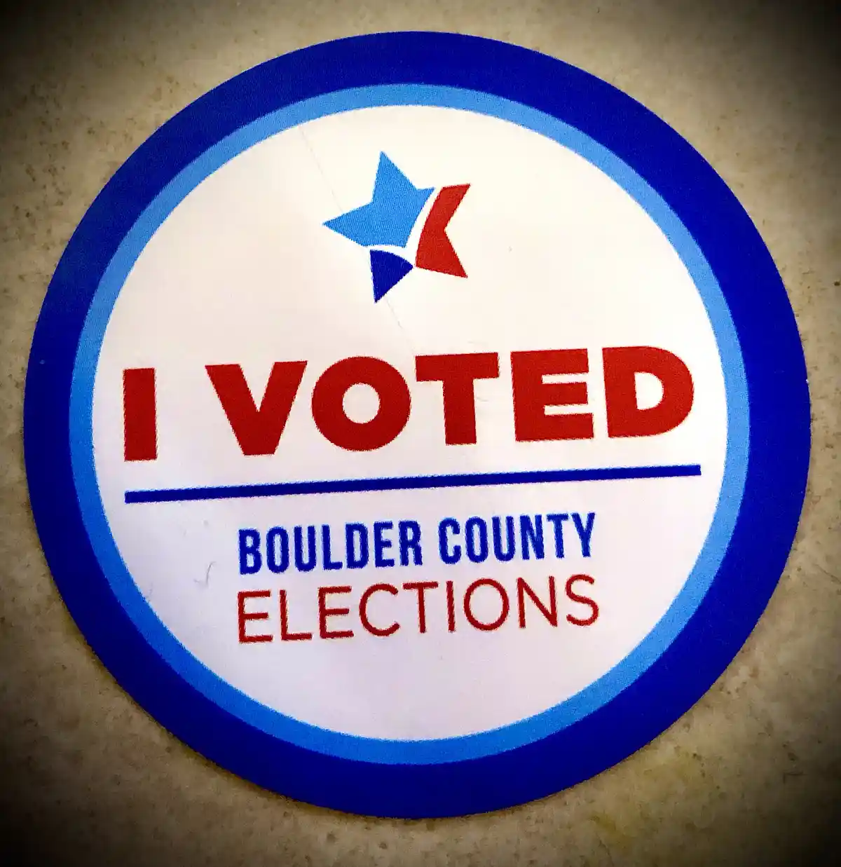 A close-up photo of a circular 'I VOTED' sticker from Boulder County Elections, featuring a blue and red star logo, red text…