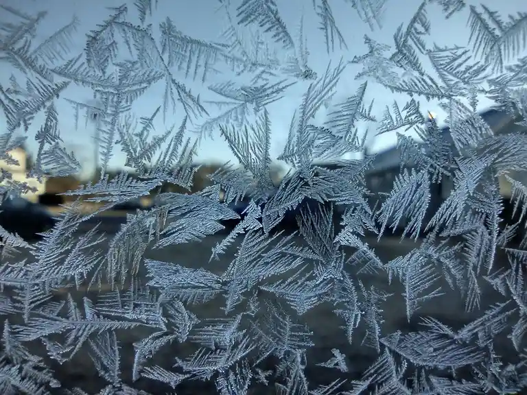 A close-up of intricate frost patterns resembling snowflakes covering a glass surface, with a blurred outdoor scene visible…