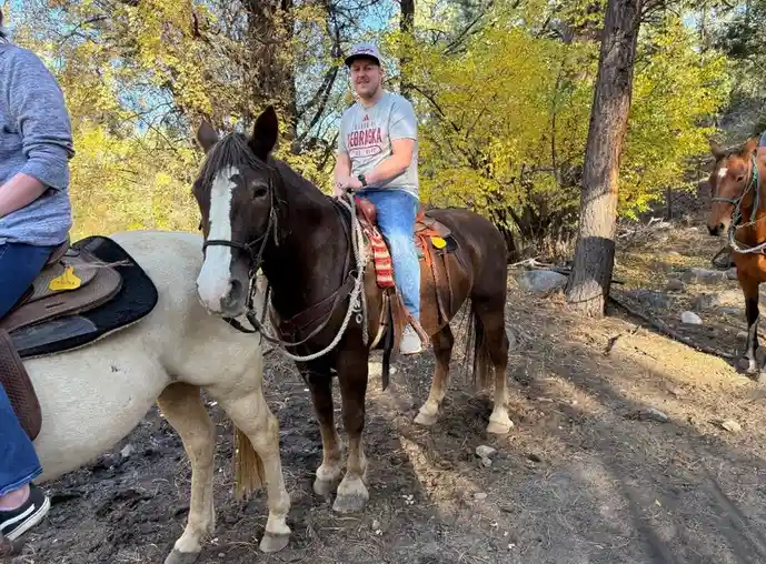 A man in a Nebraska t-shirt and baseball cap sits atop a brown horse with a white blaze, smiling at the camera. Another…