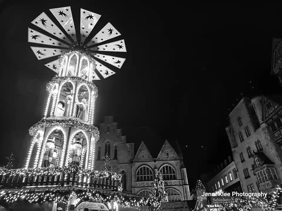 A black and white nighttime photo shows a brightly lit, multi-tiered Christmas pyramid with star-patterned blades at the…