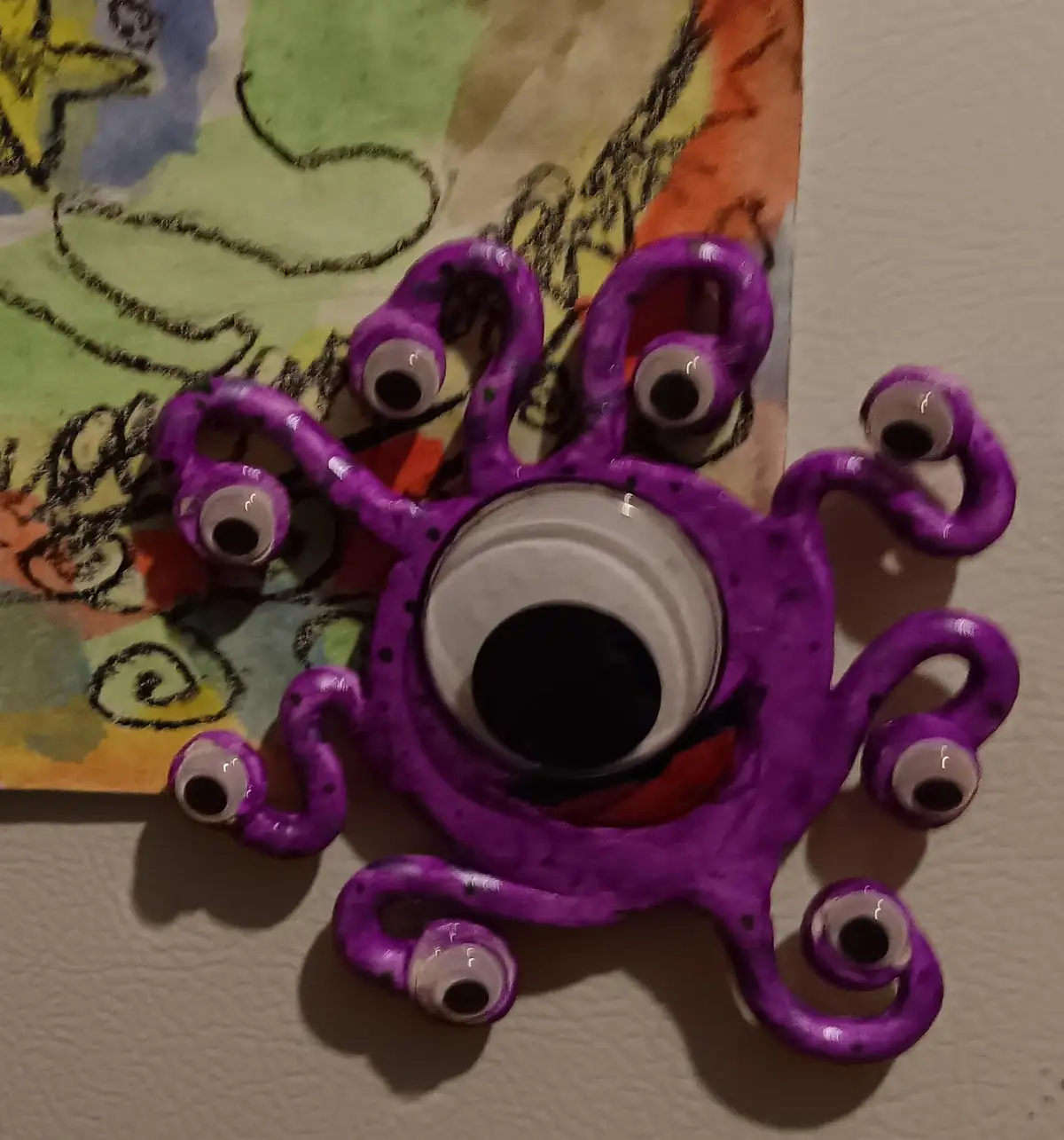 A handmade purple clay monster with one large central eye and eight smaller eyes on its tentacles, resting on a colorful…