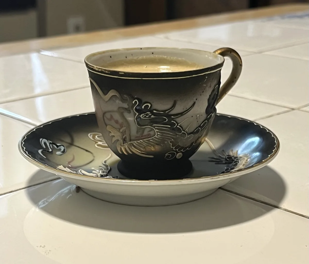 A close-up photo shows an ornate, dark-colored ceramic tea cup with a gold handle and matching saucer, both decorated with…