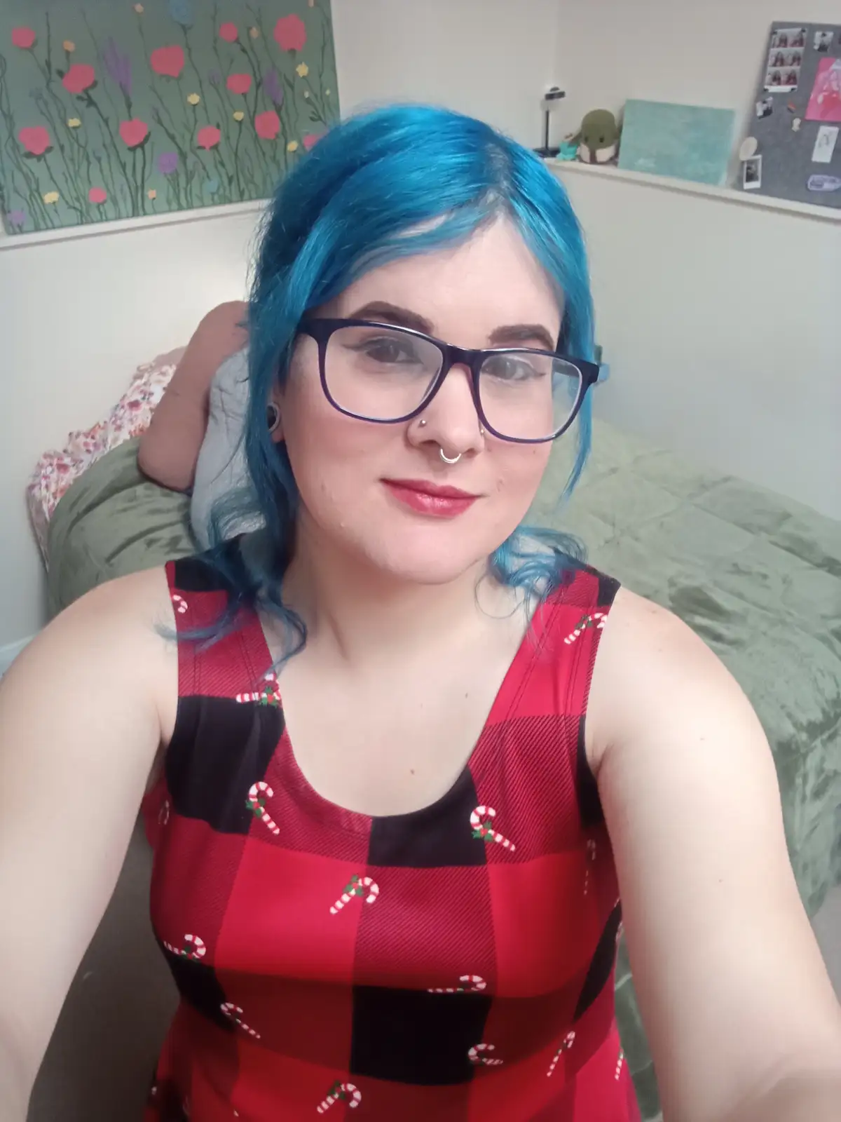 A person with bright blue hair and glasses sits on a bed wearing a red and black plaid dress decorated with candy cane…