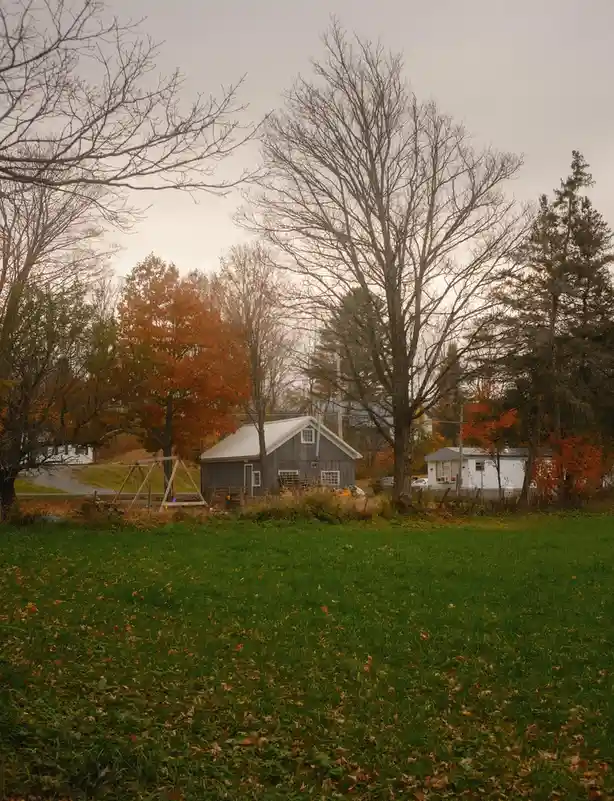 A rural autumn scene with a grassy field in the foreground, bare trees, and a small gray house with a white roof. A swing…