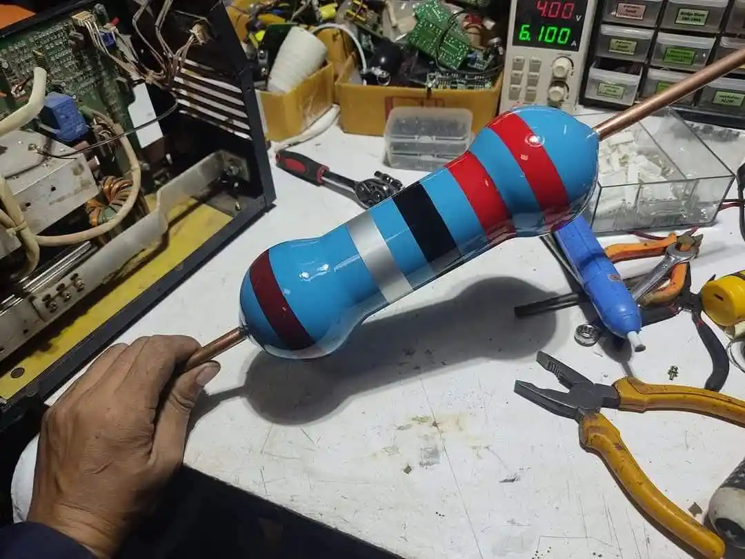 A hand holds a custom-made, colorful resistor with blue, red, and black stripes, shaped like a dumbbell, in a cluttered…