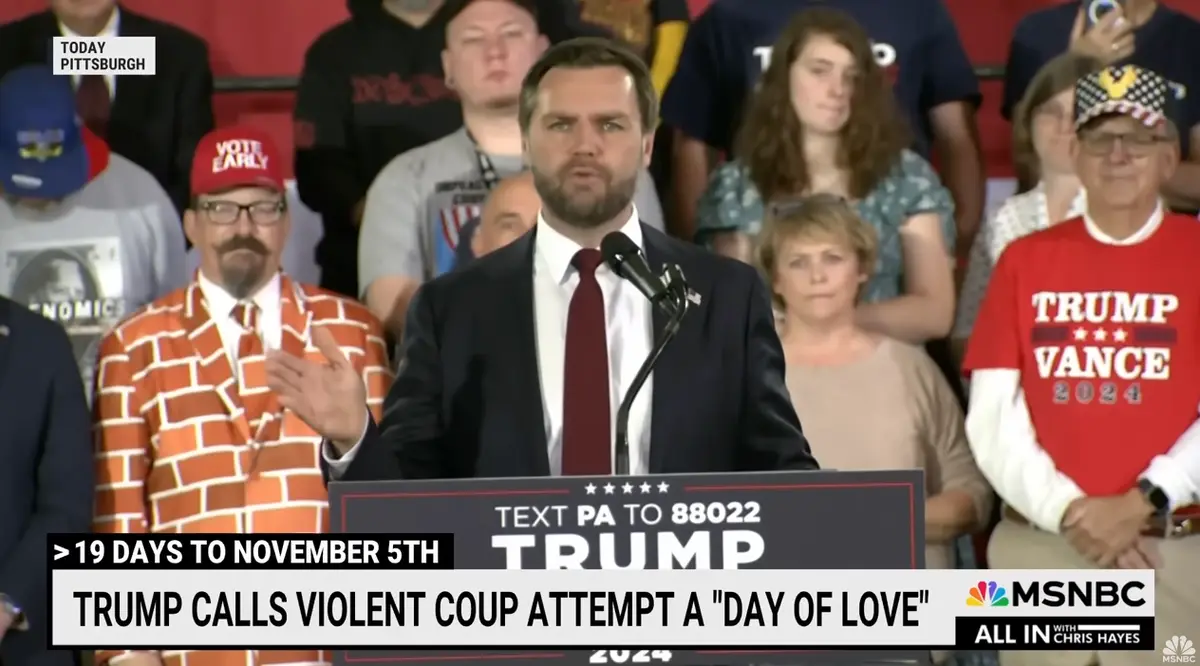 A man in a suit and red tie speaks at a podium with 'TRUMP 2024' and 'TEXT PA TO 88022' visible. Behind him, a crowd…