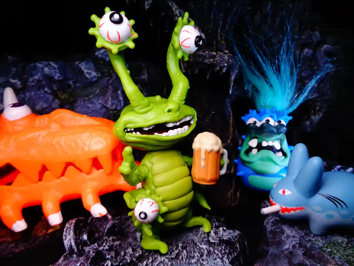 A green toy monster with three eyeballs on stalks, holding a beer mug and giving a thumbs-up, stands among other colorful…