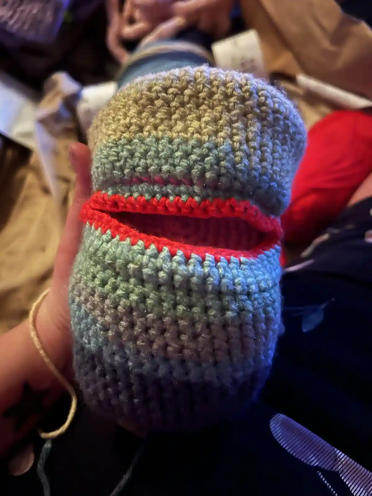 A hand holds a crocheted baby bootie that has been humorously altered to resemble a wide, open mouth with red yarn stitching…