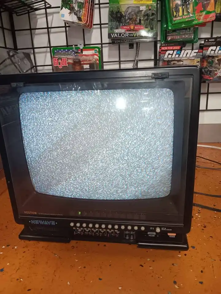 An old black Veston Newave CRT television sits on an orange floor, its screen filled with static noise, with toy action…