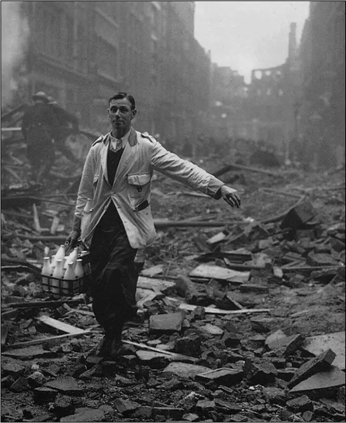 A black-and-white historical photo shows a man in a light-colored trench coat walking through rubble-strewn ruins, carrying…