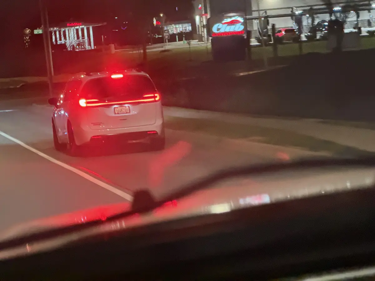 A nighttime photo taken from inside a car shows a white SUV with its brake lights on, driving away from a 'Club Car Wash'…