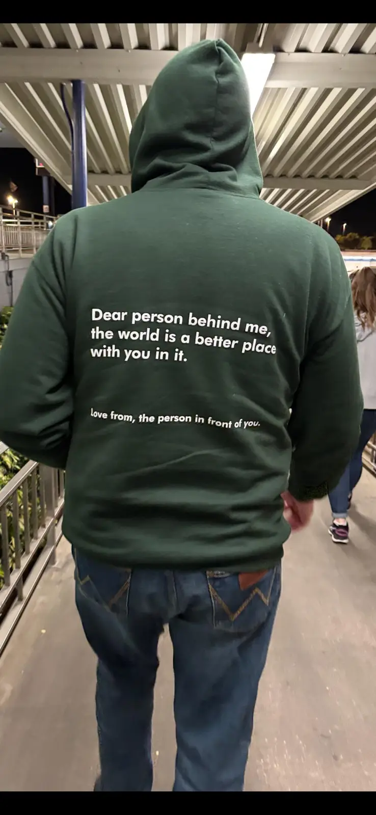 A person wearing a dark green hoodie with white text on the back walks away from the camera on an outdoor walkway at night…