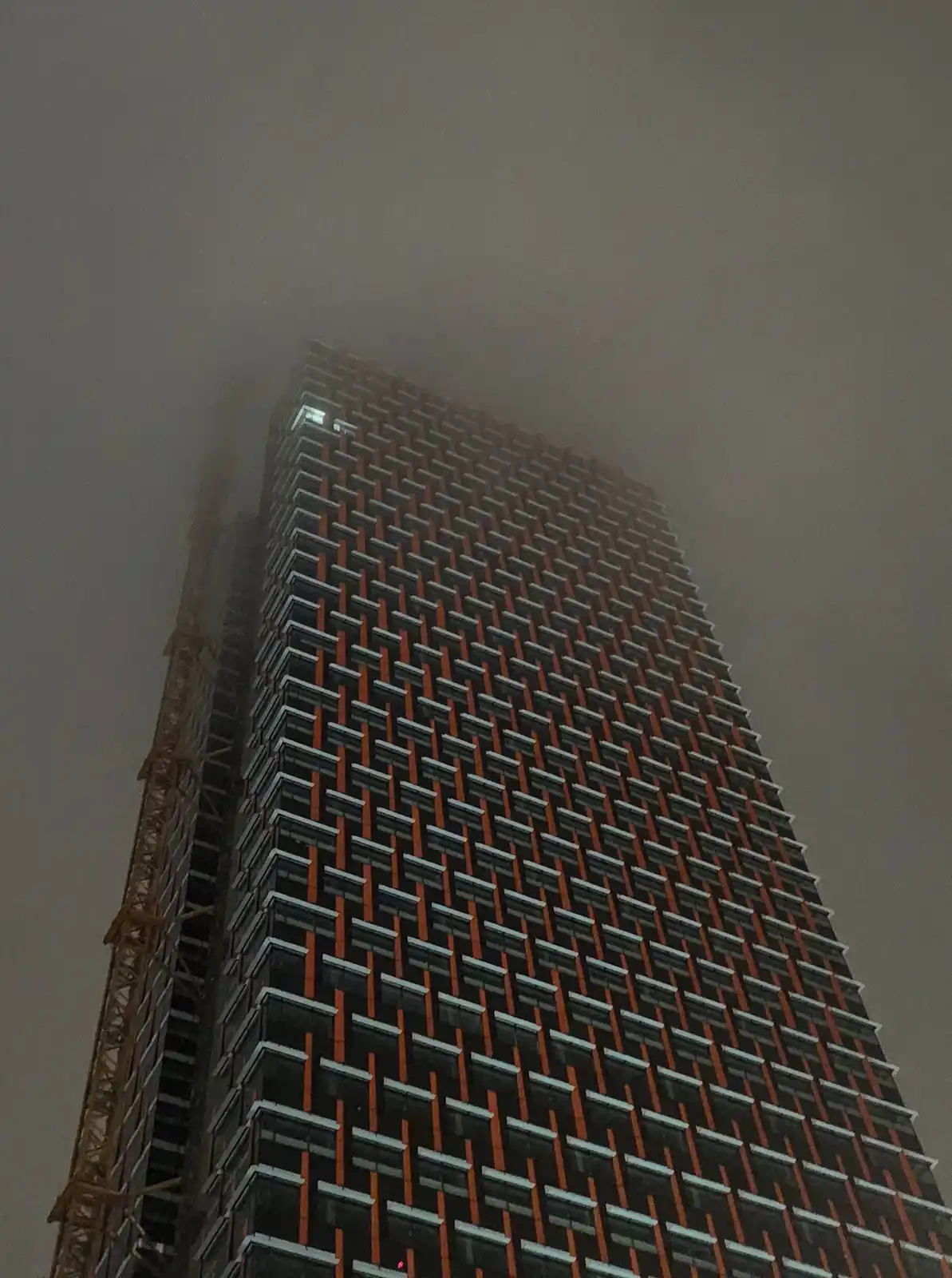 A low-angle shot of a tall skyscraper partially obscured by thick fog, with a construction crane visible on its left side…