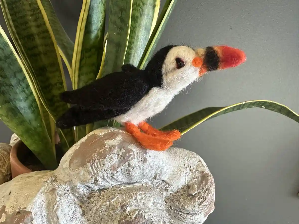 A felt puffin figurine with black and white feathers, orange beak and feet, perched on a weathered stone statue, positioned…