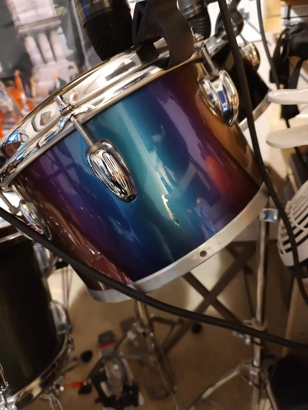 A close-up photo of a shiny, multi-colored drum with chrome hardware, a microphone mounted above it, and cables running…