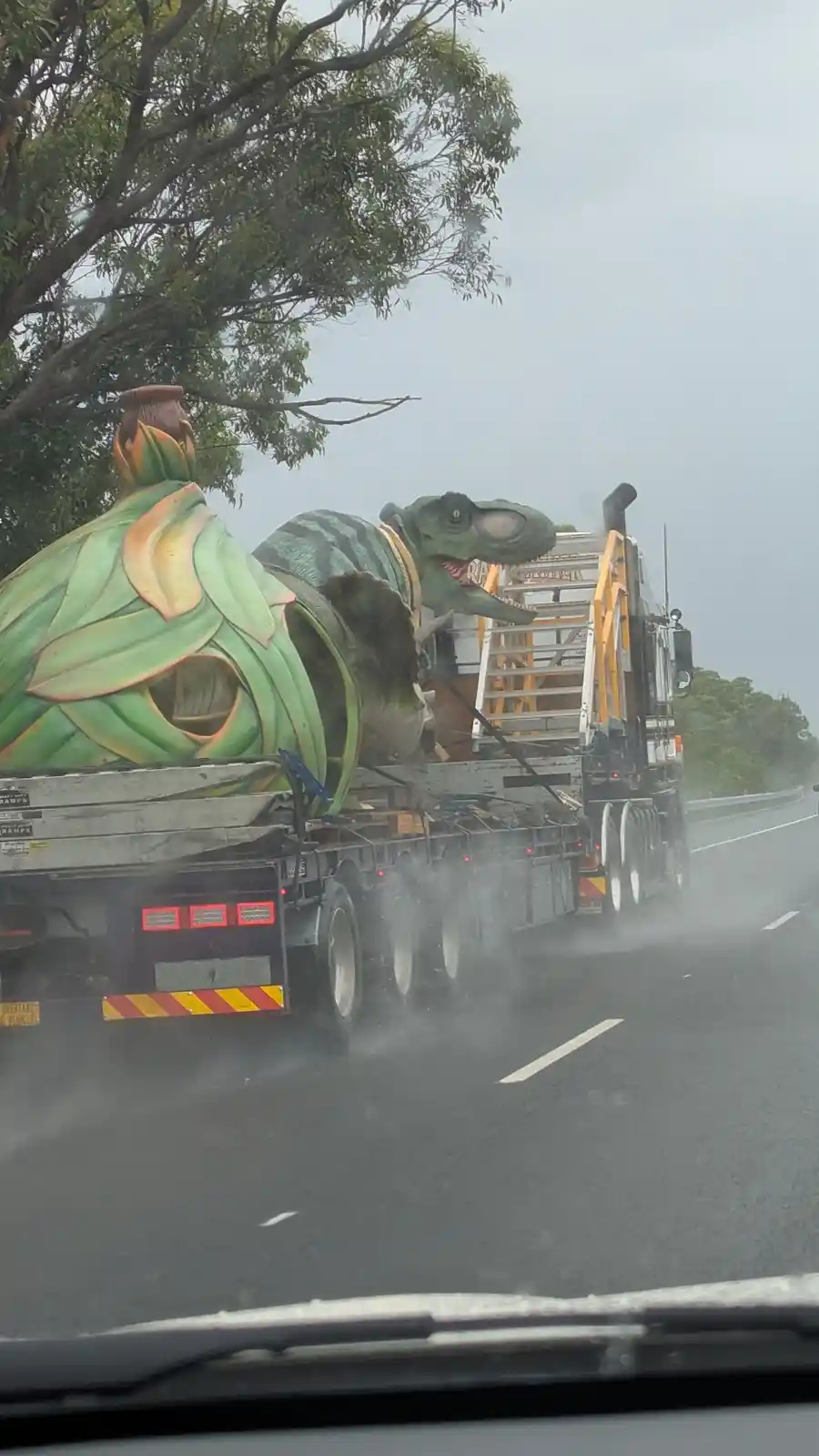 A large truck is hauling oversized, colorful dinosaur and plant-themed ride-on attraction props on a wet highway, viewed…