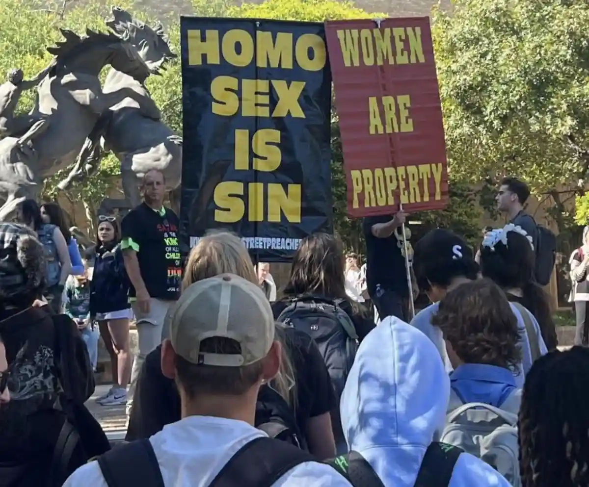 A crowd of people outdoors, some facing away, with two large protest signs visible: one black with yellow text reading 'HOMO…