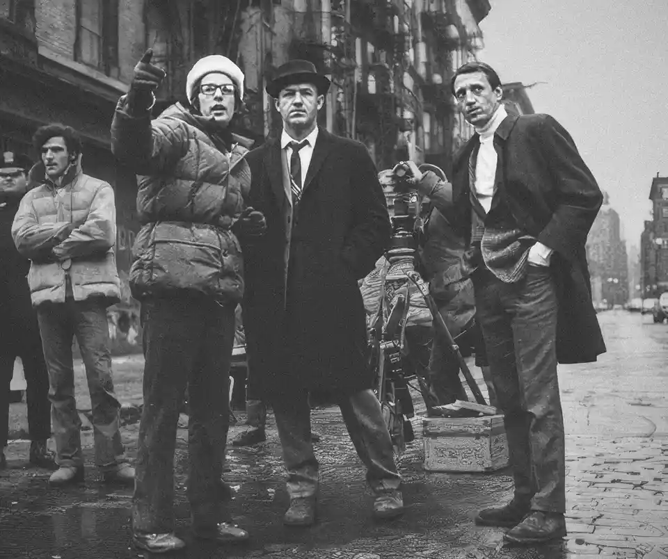 A black-and-white photo shows a group of men on a wet city street, one pointing emphatically while another stands beside a…