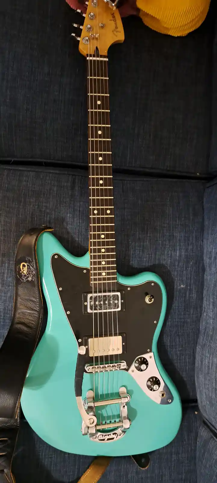 A turquoise Fender Jaguar electric guitar with a black pickguard and chrome hardware rests on a dark blue denim couch, with…