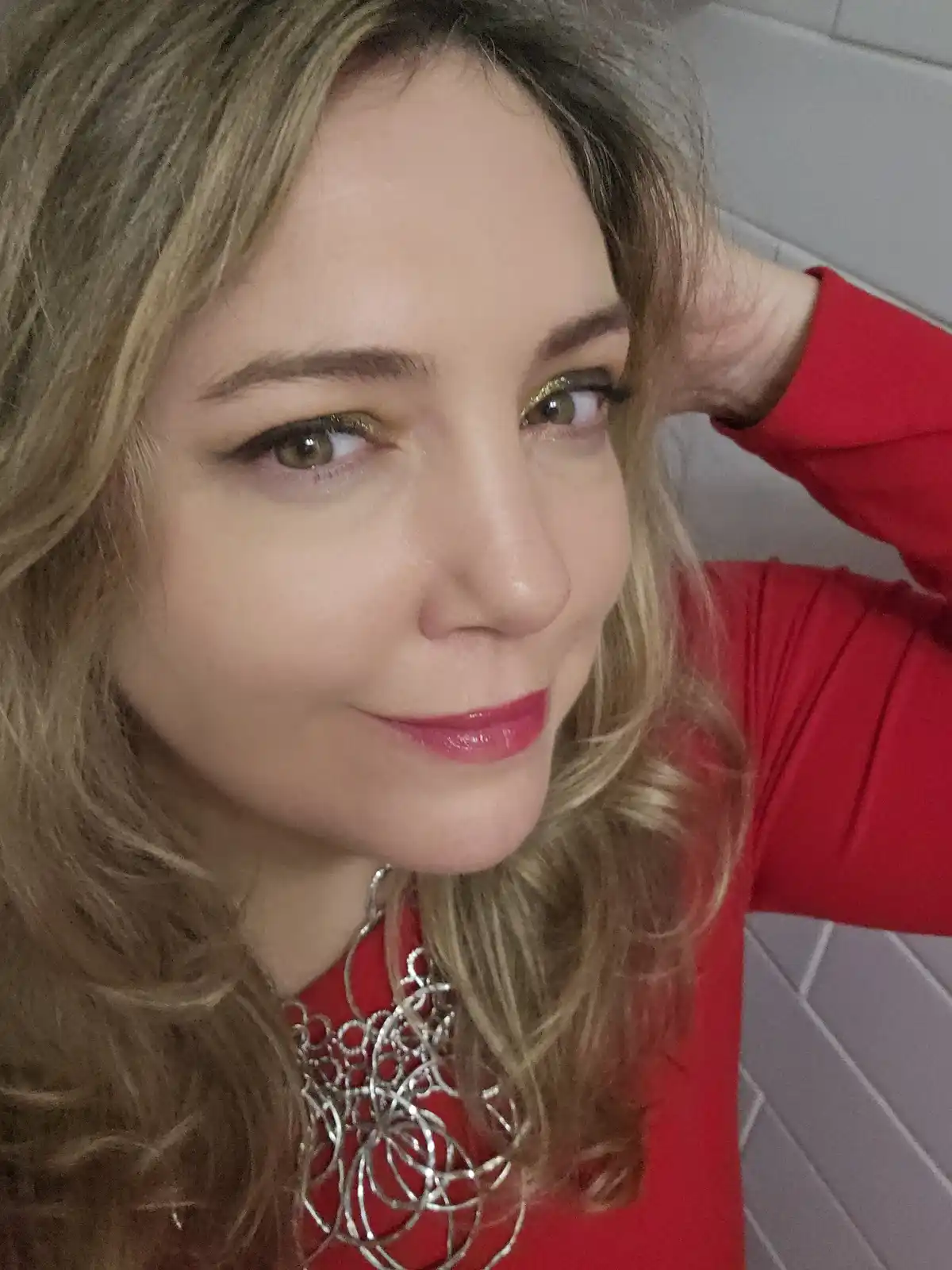 A woman with blonde hair and glittery gold eyeshadow looks at the camera with a slight smile while wearing a red top and a…