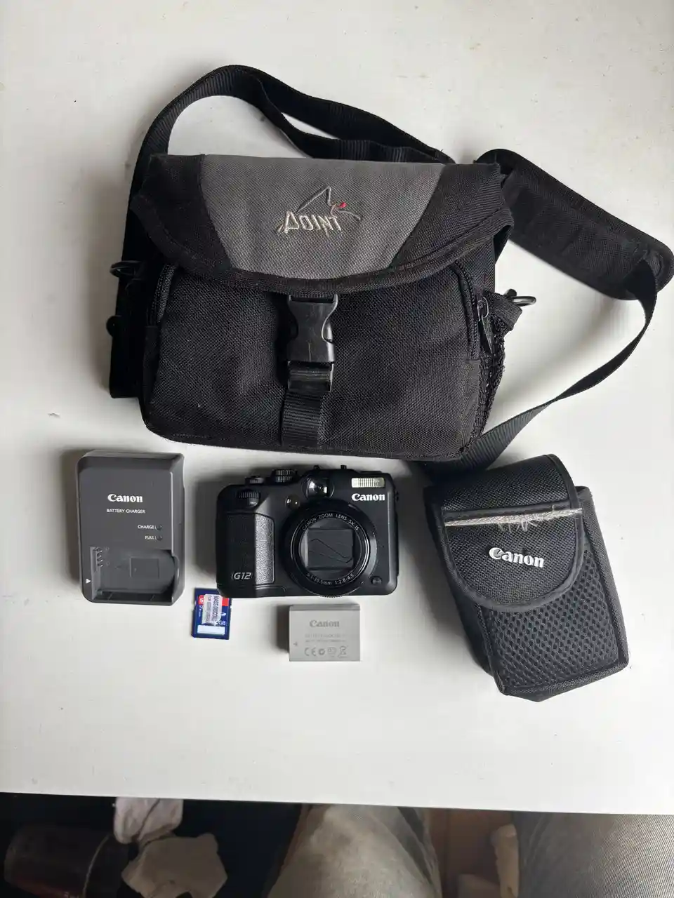 An overhead shot displays a Canon G12 point-and-shoot camera, its matching battery, charger, memory card, and protective…