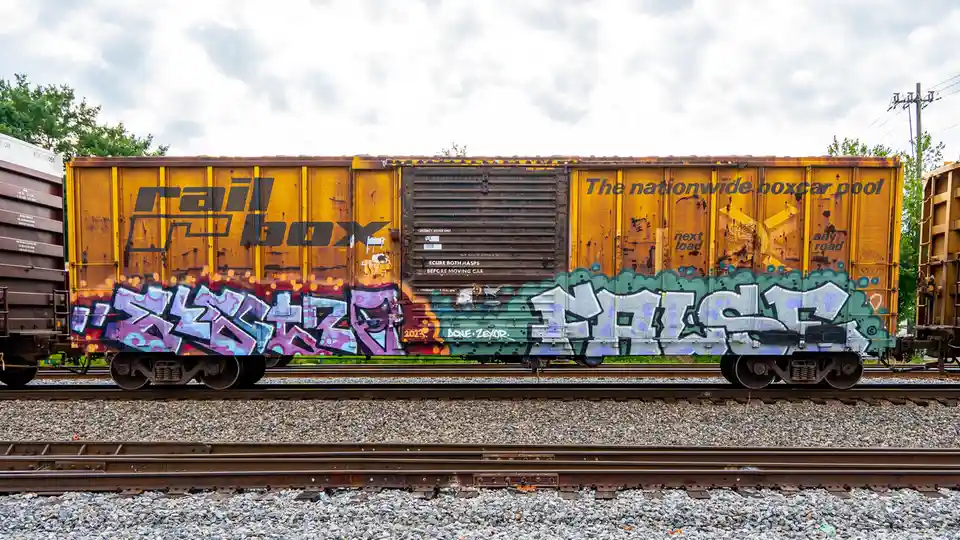 A yellow railroad boxcar with 'rail box' and 'The nationwide boxcar pool' printed on it, covered in colorful graffiti, sits…