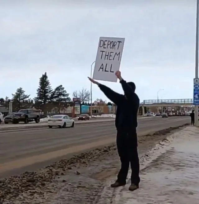 A person wearing dark clothing stands on a snowy roadside holding up a white sign with bold black handwritten text that…