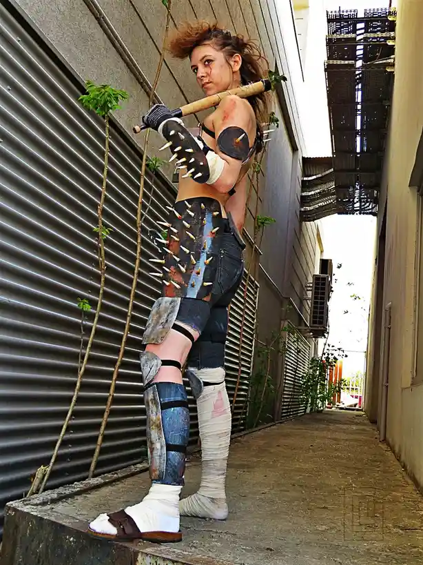 A woman in a makeshift, heavily spiked and bandaged warrior costume stands in an alleyway, holding a wooden bat with spikes…