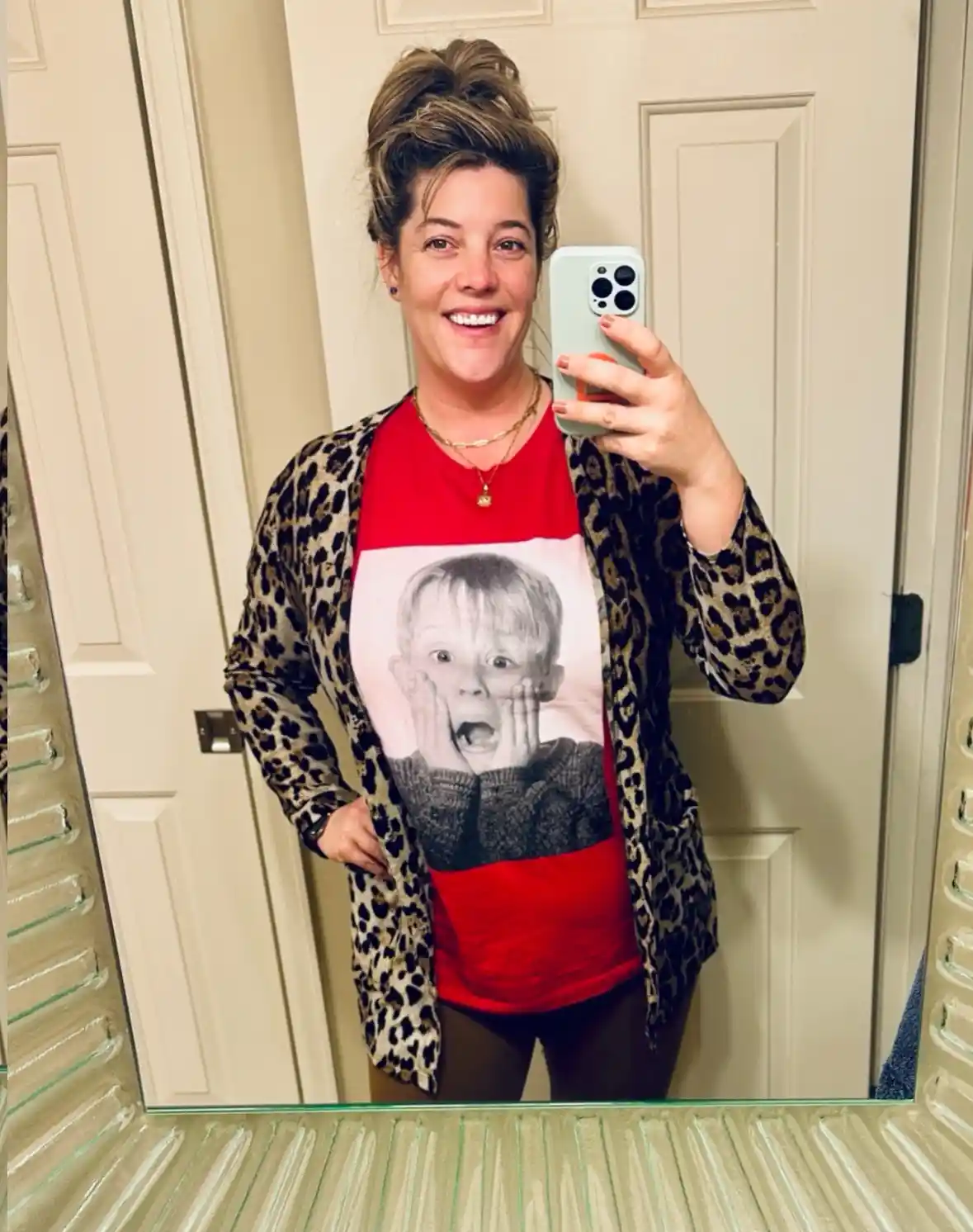 A woman takes a mirror selfie wearing a red t-shirt with a large black-and-white image of a young boy (Macaulay Culkin as…