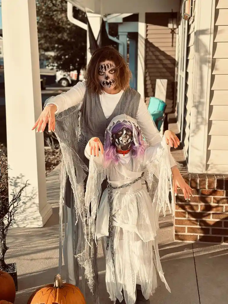 Two people, an adult and a child, are dressed in elaborate zombie costumes on a porch. The adult has skull makeup and wears…