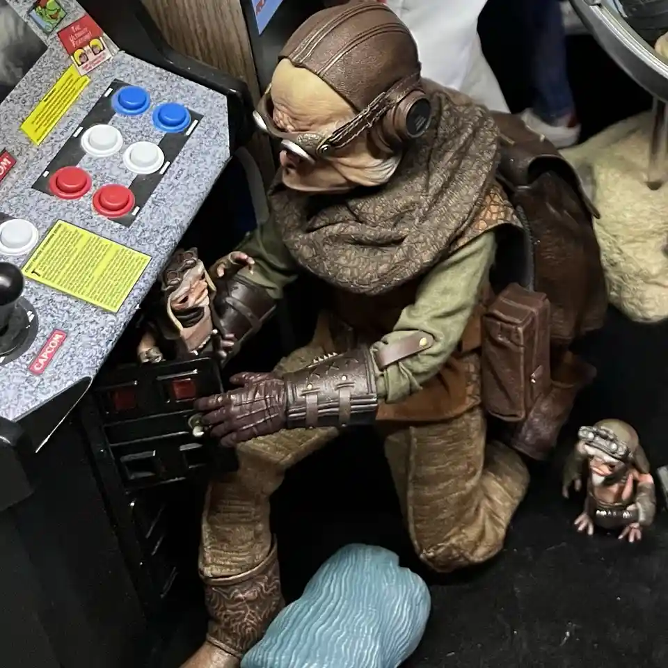 A person in a detailed, brown leather and fabric costume resembling a Star Wars character kneels before a vintage arcade…