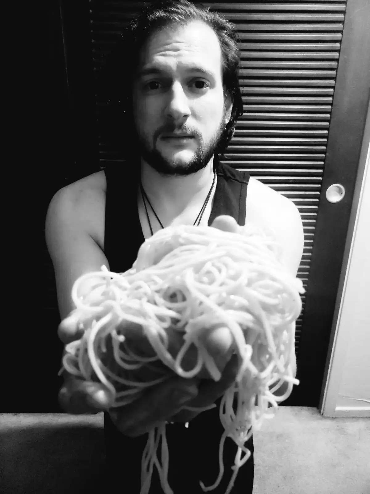 A black-and-white photo shows a man with a beard and tank top holding a large, messy pile of cooked spaghetti noodles…