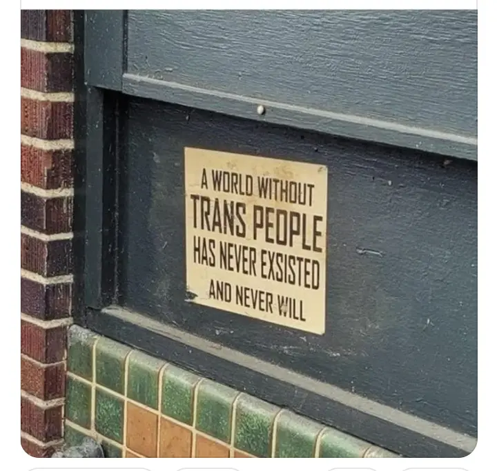 A close-up photo shows a beige rectangular sign affixed to a dark gray wooden door frame next to a brick wall and green…