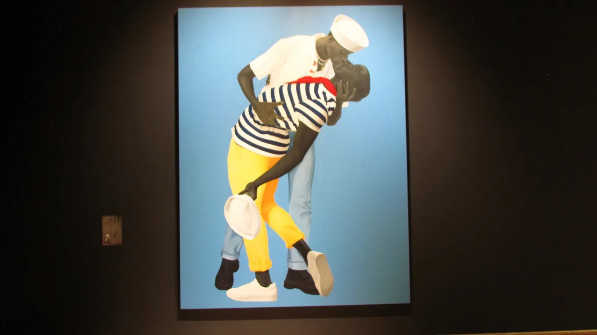 A framed photograph on a dark wall shows two Black men in sailor-style outfits embracing passionately against a solid blue…