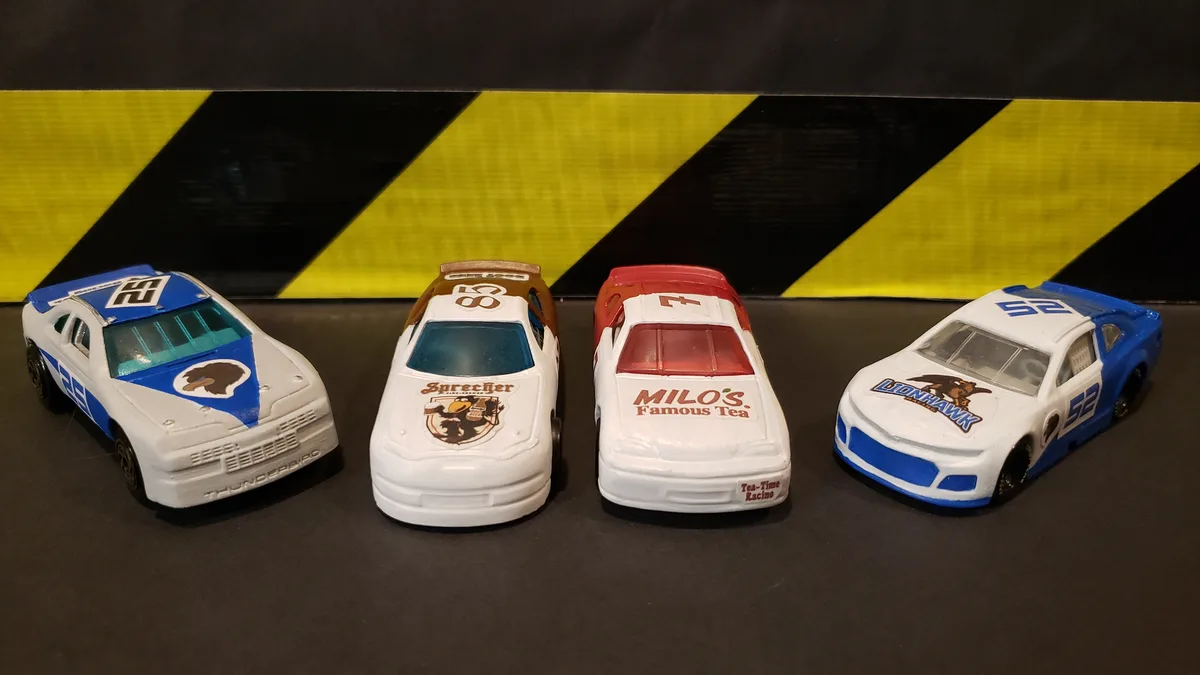 Four toy race cars are lined up in front of a black and yellow hazard stripe background. Each car has a different sponsor…