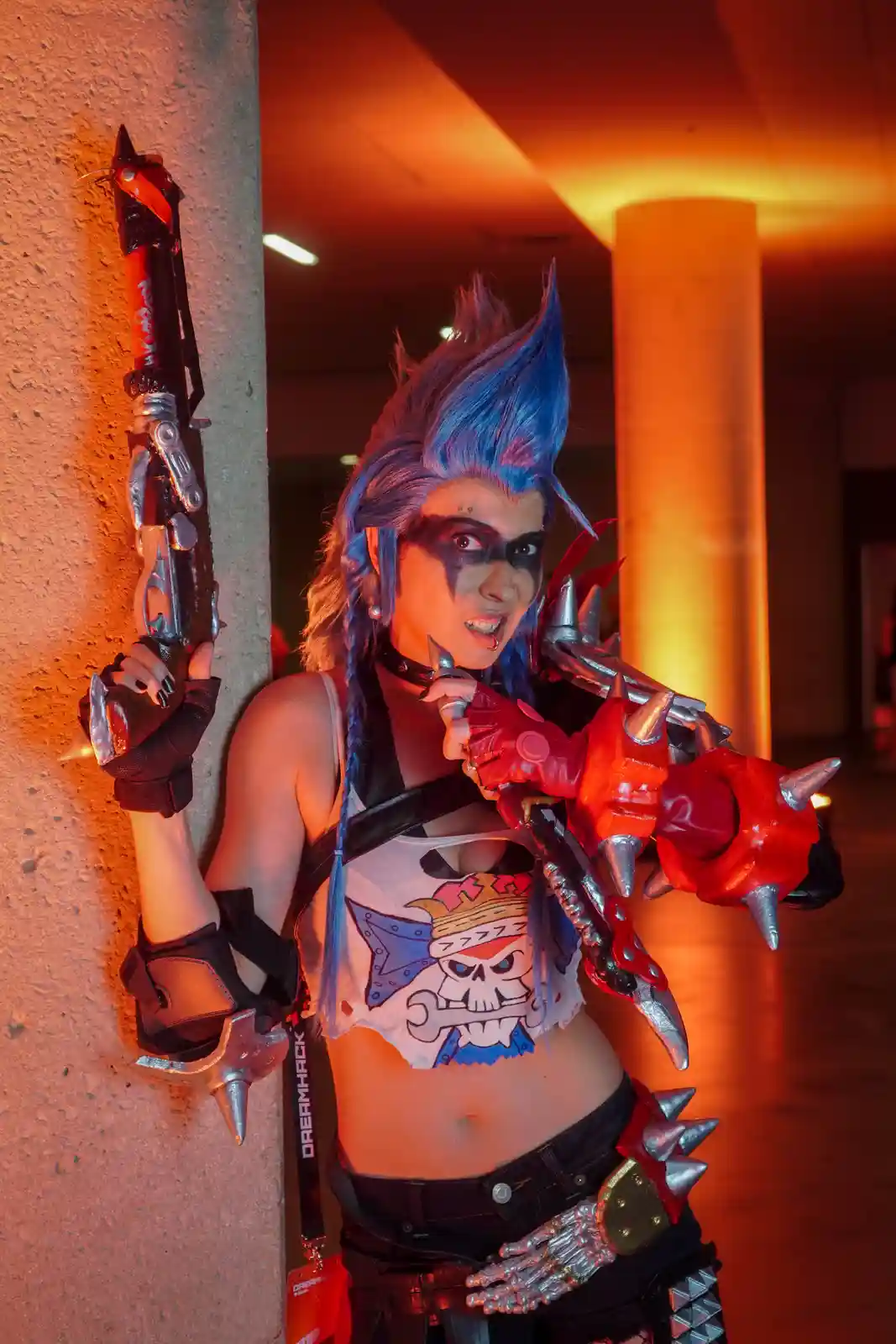 A person in elaborate cosplay with blue spiky hair, black eye makeup, and multiple weapon props leans against a wall. They…